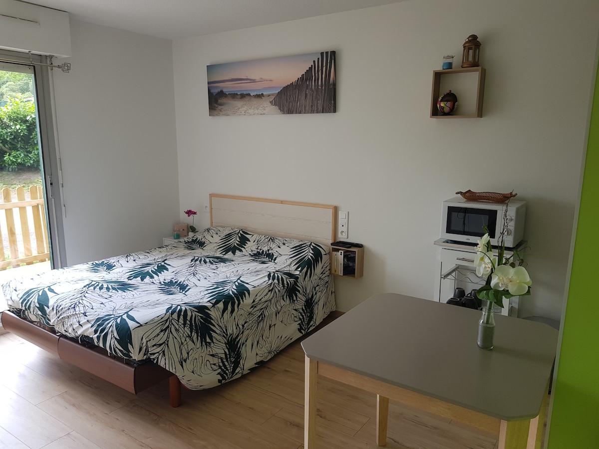 A well-lit studio space features a spacious bed adorned with a leafy patterned comforter. A small dining table is positioned nearby, accompanied by a vase with fresh flowers. A microwave and decorative shelves are evident, while large glass doors offer access to the exterior.