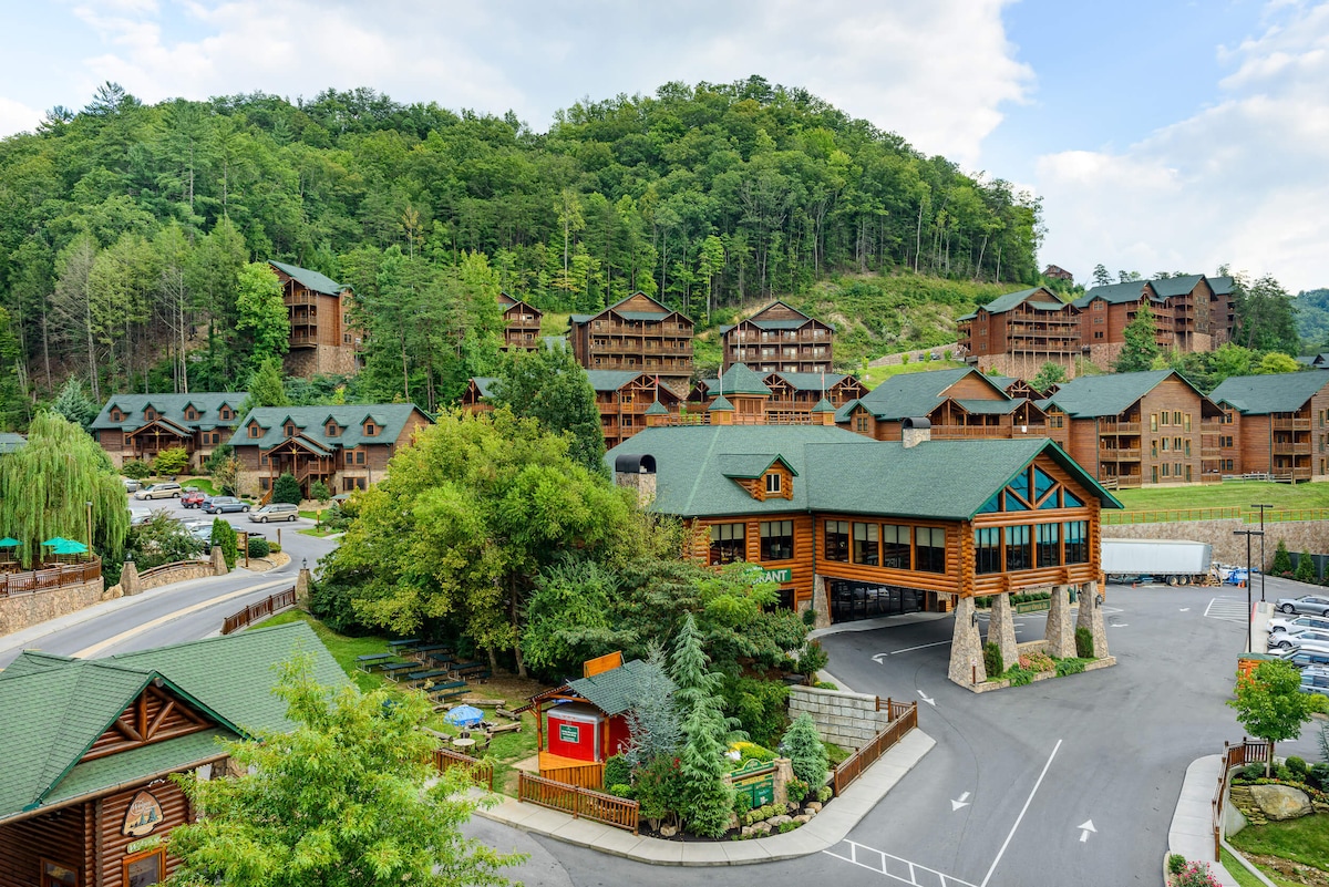 A view of the resort complex is presented, featuring multiple wooden buildings nestled among lush greenery. The sloped rooftops are covered with dark shingles, while the surrounding hillside is filled with trees, creating a natural backdrop. A clear blue sky enhances the serene setting.