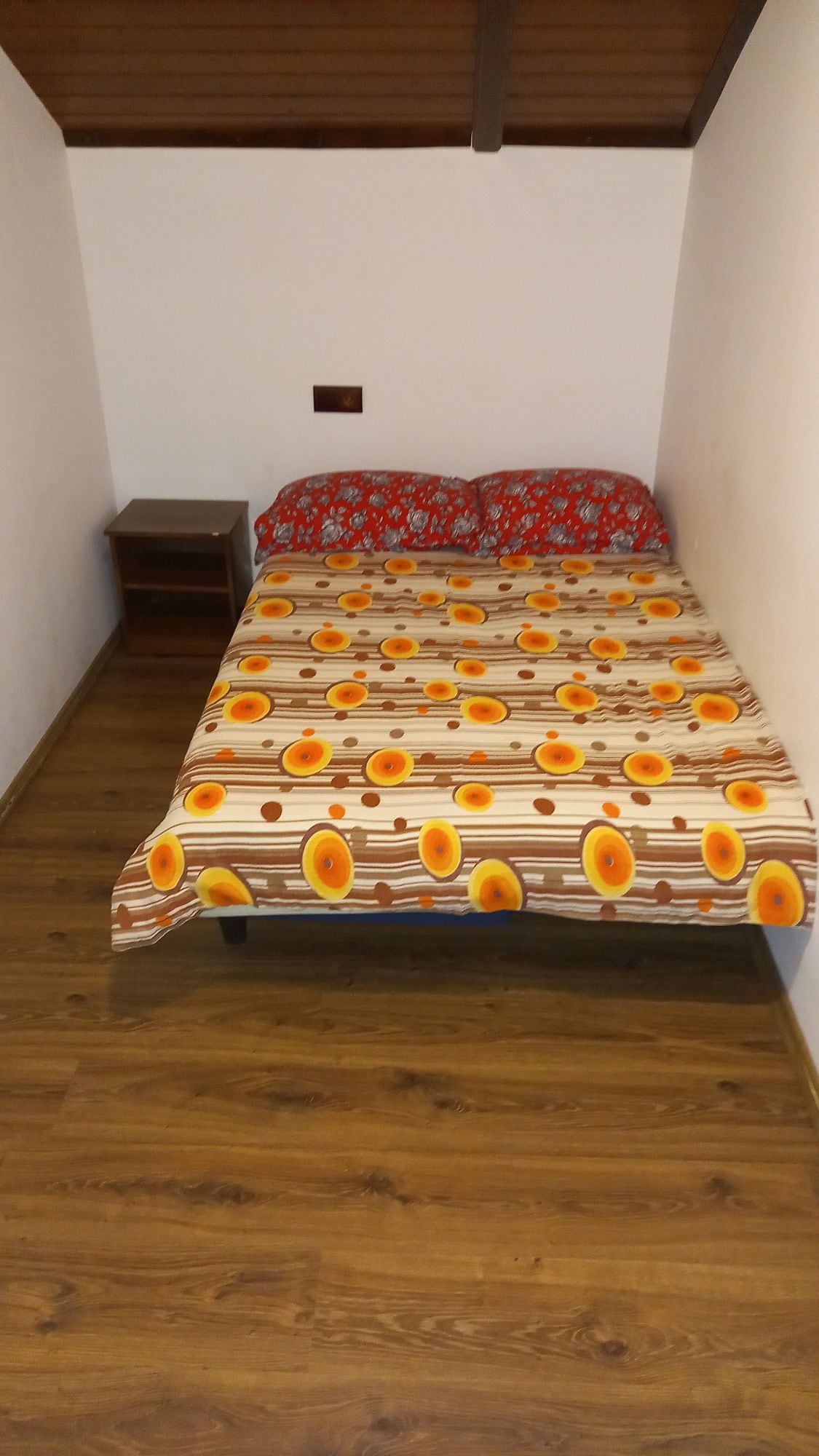 A sleeping area features a spacious bed draped with a floral-patterned blanket in shades of orange and red. A bedside table is positioned next to the bed, and warm wooden flooring complements the overall simplicity of the room.