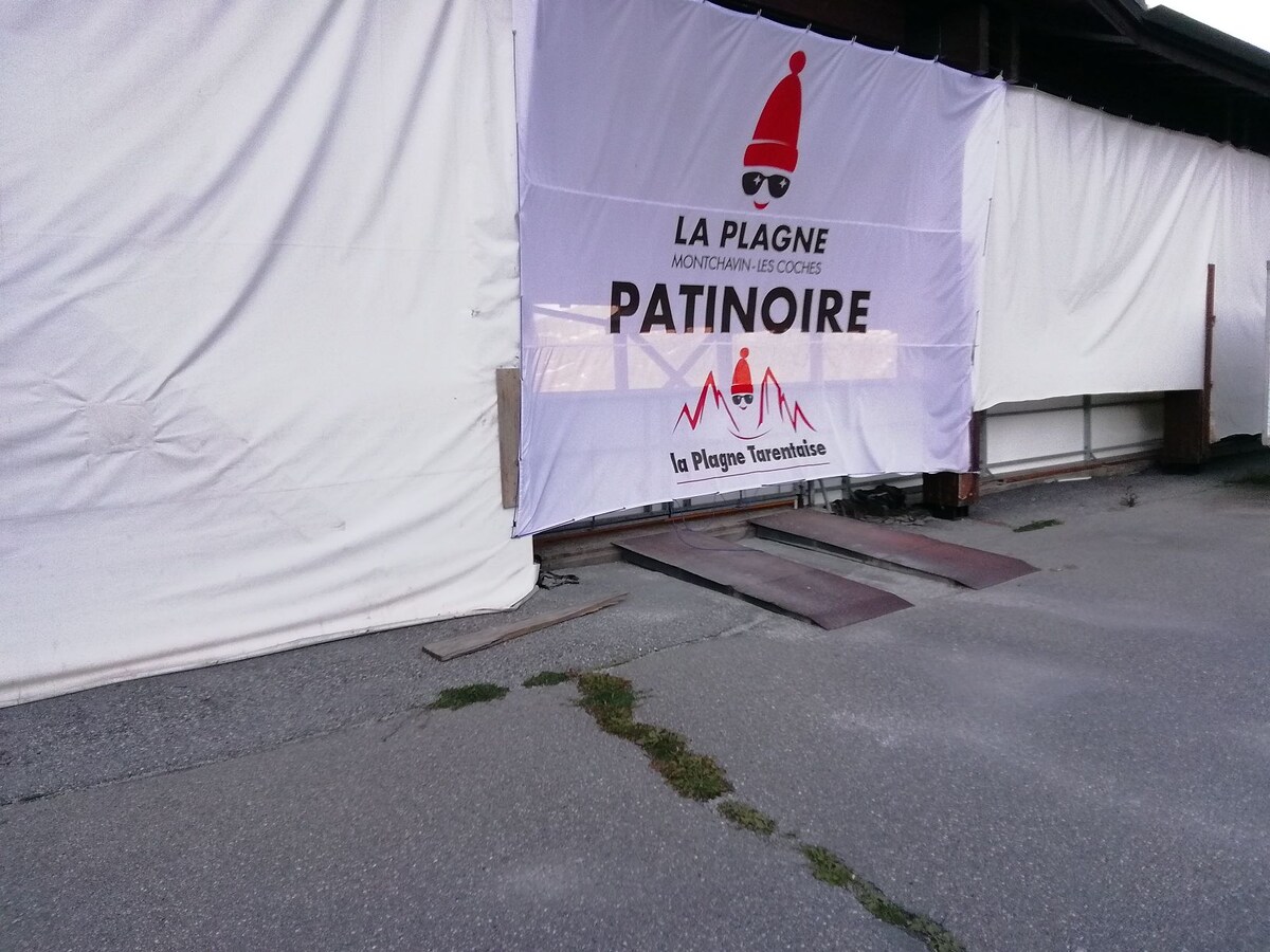 A large banner is displayed at the entrance to an ice skating rink, featuring the words 'La Plagne Montchavin-Les Coches Patinoire.' The area is paved, with a slight ramp leading into the rink, allowing accessibility for visitors.