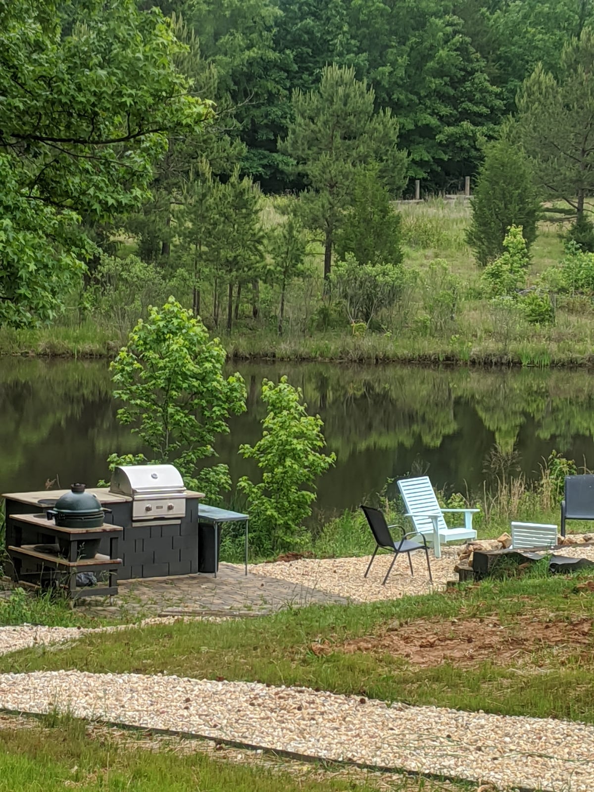 A serene outdoor space features a grilling area with a barbecue and a green egg smoker, surrounded by lush greenery. A seating arrangement with several chairs is positioned near a calm pond, reflecting the natural scenery. A gravel pathway leads through the area.