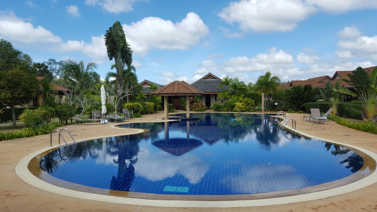An inviting communal pool features a rounded shape with vibrant blue tiles reflecting the sky above. Surrounding greenery and palm trees enhance the serene environment. Comfortable lounge chairs are placed nearby, offering a place to relax. A shaded area is visible at one corner of the pool.