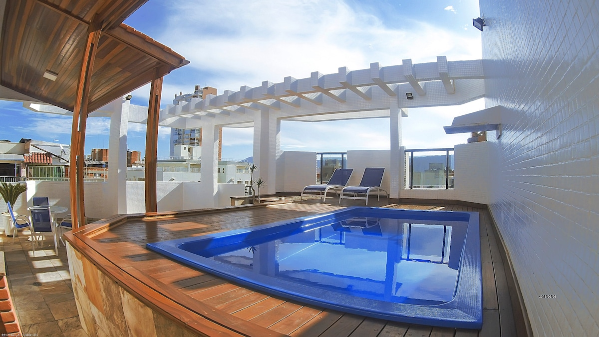 A private pool is bordered by a spacious wooden deck, featuring lounge chairs for relaxation. An overhead pergola provides shade, while large windows invite natural light into the area. The surrounding walls are finished in a clean white for a modern aesthetic.