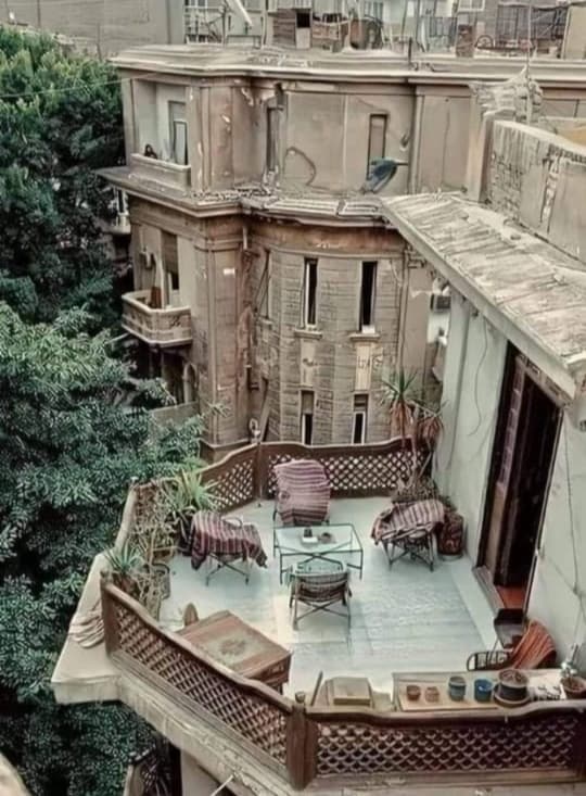 A covered terrace is adorned with plants and features a seating area, including several chairs and a glass table. The terrace offers views of surrounding greenery, creating a tranquil outdoor retreat within the urban environment.