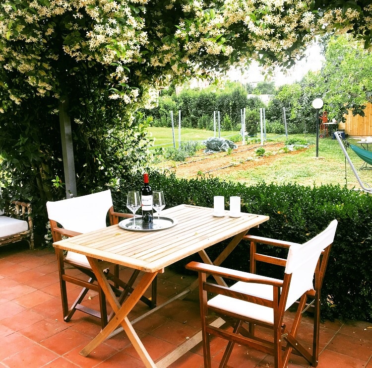 A wooden dining table is set with a bottle of wine and glasses, surrounded by two white cushioned chairs. Lush greenery frames the outdoor space, with a garden visible in the background, showcasing a variety of plants and serene landscaping.