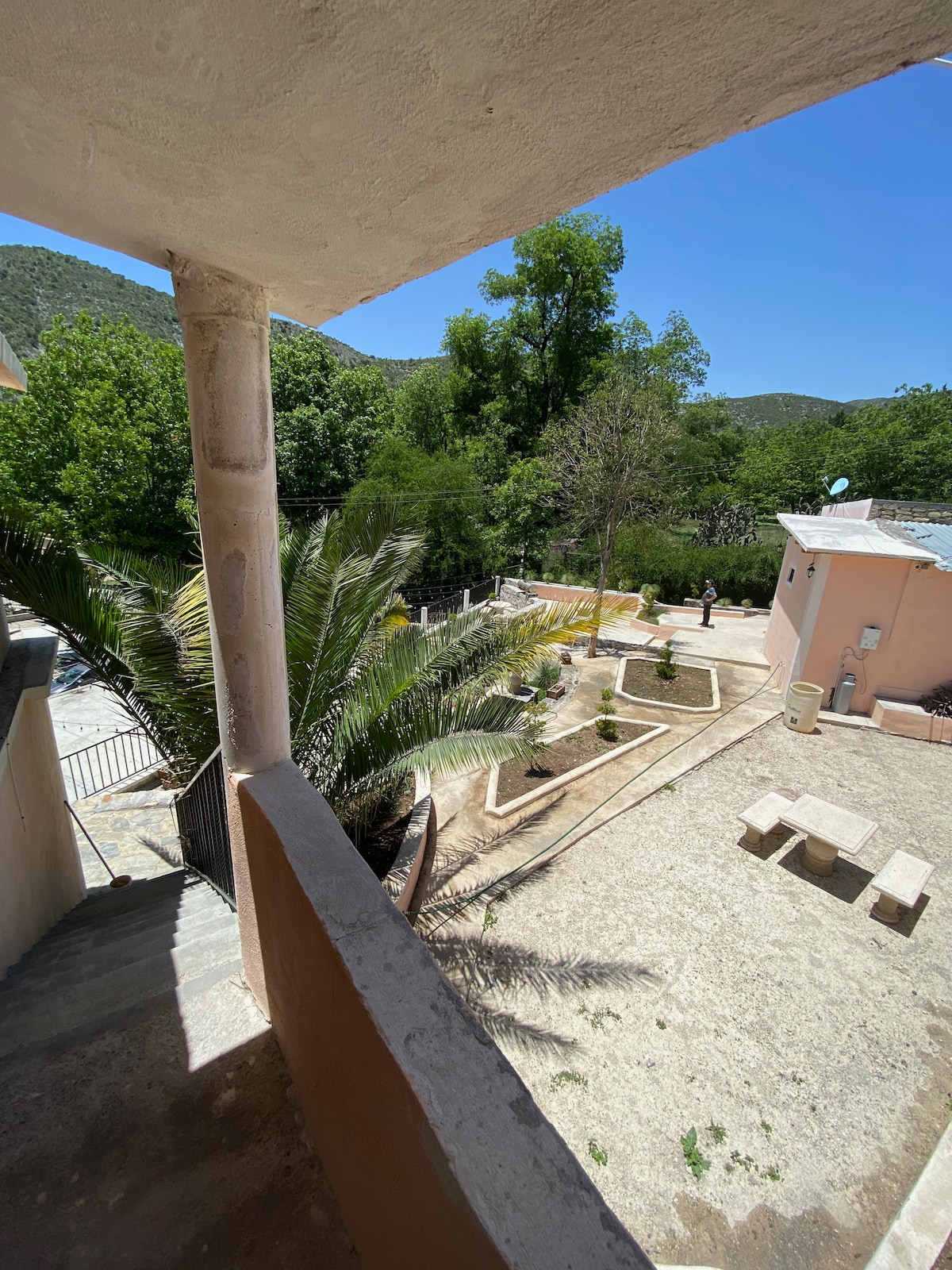 A view from the balcony reveals a landscaped outdoor area with stone pathways and sections of garden. Lush green trees and shrubs surround the space, providing a natural setting. In the distance, a person can be seen walking along the pathway, contributing to the serene atmosphere.