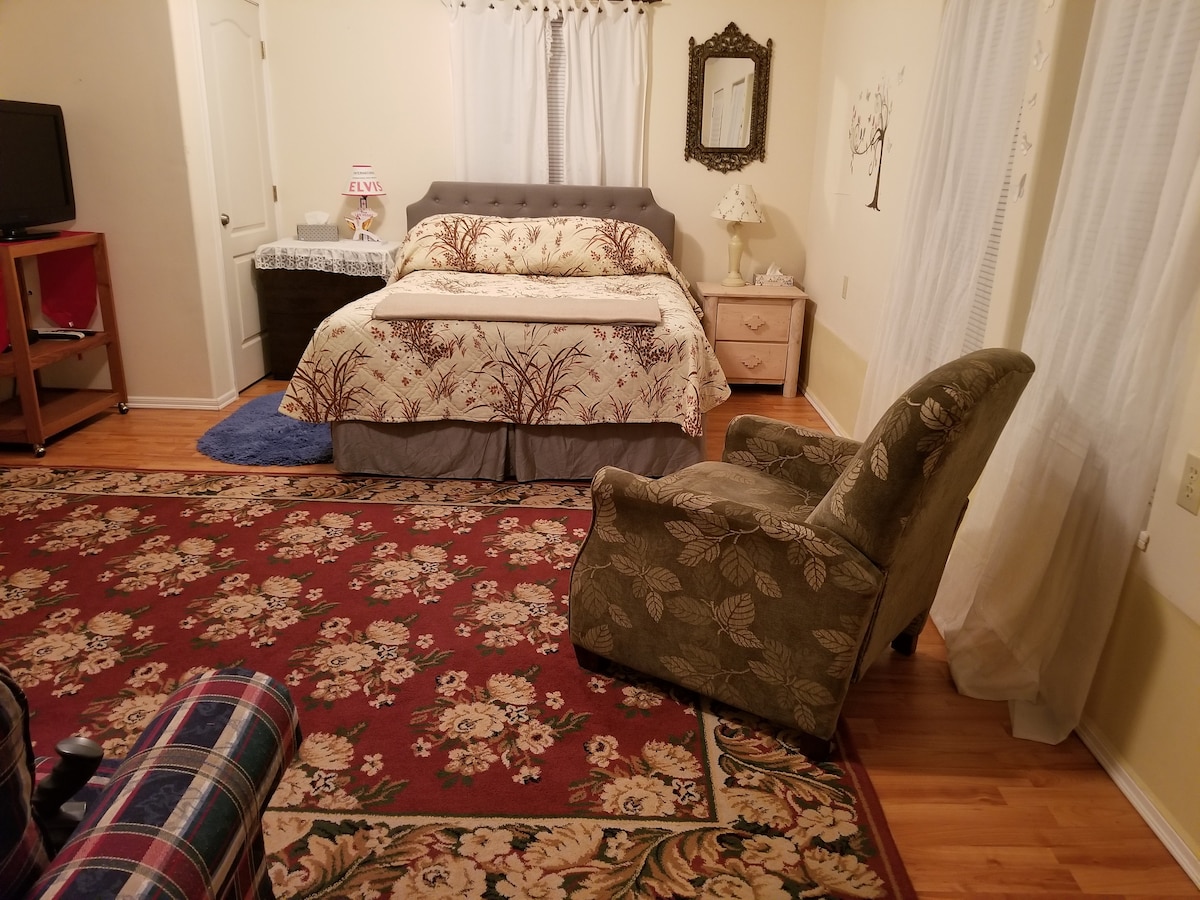 A cozy room features a queen bed with a floral coverlet and a comfortably positioned armchair. A patterned area rug adorns the wooden floor, complementing bedside tables with lamps. A small television on a wooden cart and draped curtains allow for natural light to enter.