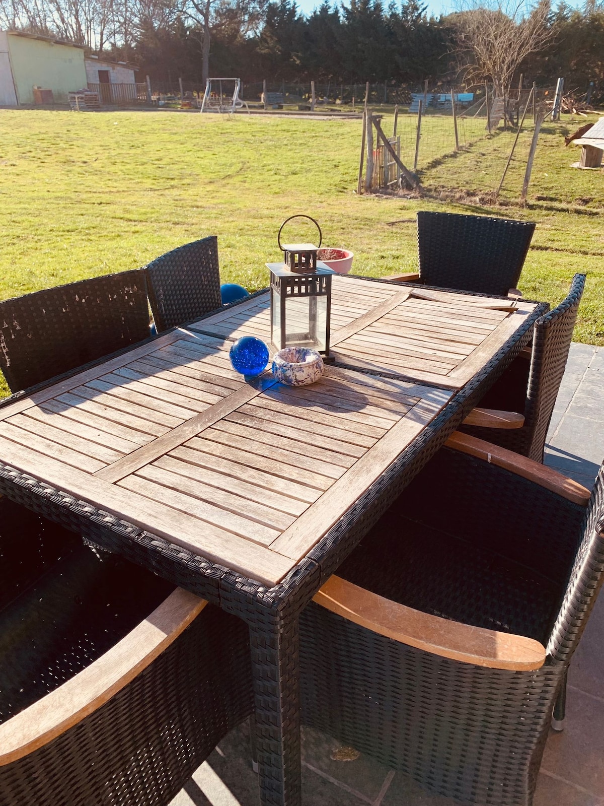 A wooden outdoor table is surrounded by six woven chairs, situated on a patio. A decorative lantern and a colorful object are placed at the center of the table. In the background, a green lawn extends towards a tree line.