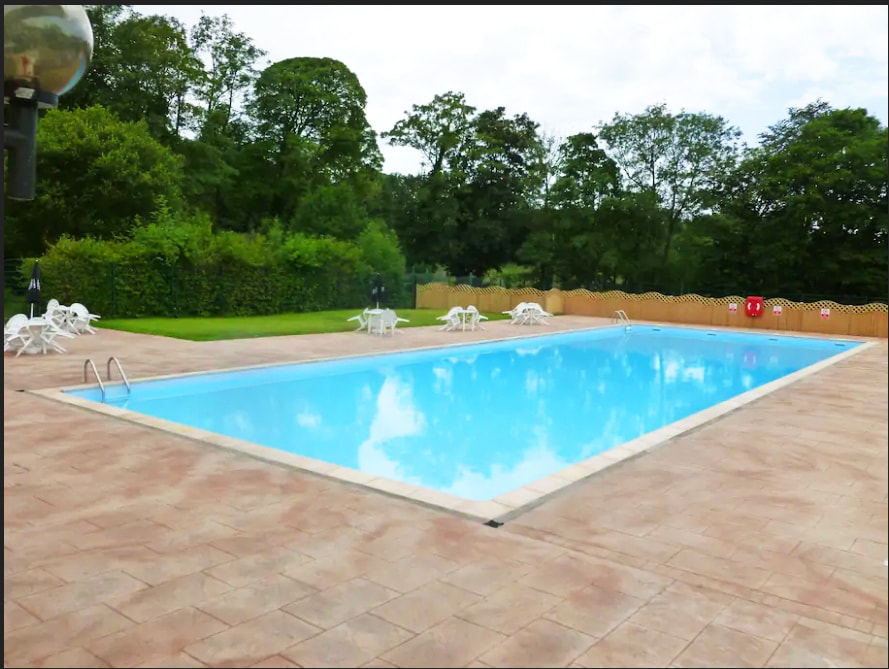 An outdoor swimming pool is set within a green landscape, featuring clear blue water reflecting the sky. Surrounding the pool are several white lounge chairs arranged on a paved deck. Lush trees frame the area, providing a peaceful atmosphere.