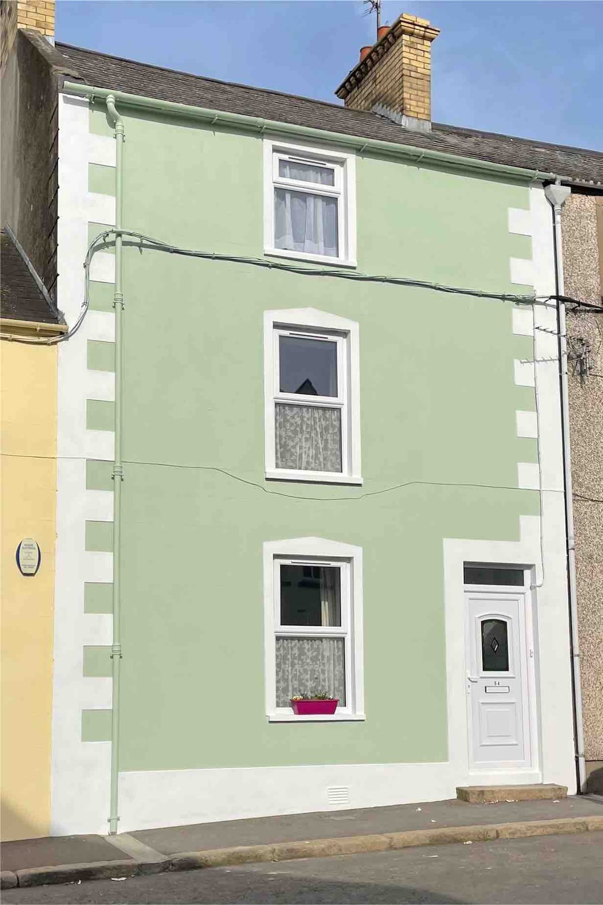 A three-story house features a light green facade complemented by white trim. Two windows, adorned with lace curtains, are positioned symmetrically on the upper floors, while a simple white door is located at the front, offering access to the residence.