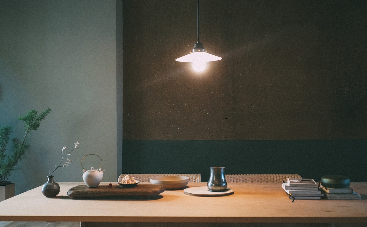 A minimalist dining table is highlighted by an overhead pendant light, casting a warm glow. Various dishes and decorative items are arranged on its surface, including a small vase with delicate branches, a teapot, and a stack of books in the background.