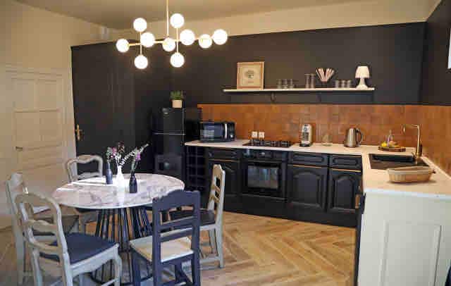 A modern kitchen is presented with dark cabinetry and warm wood accents. A round dining table with light chairs is positioned in the center. Stainless steel appliances, including a microwave and kettle, are visible, alongside a charming light fixture that illuminates the space.