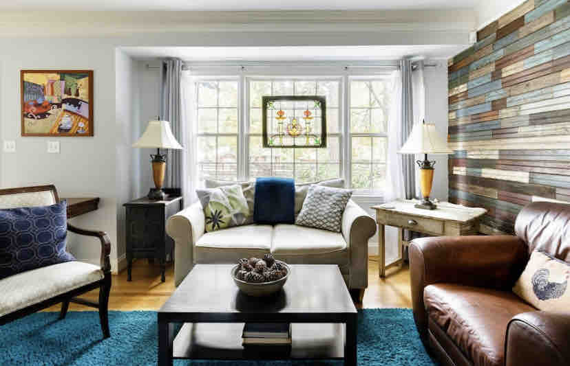 The living area presents a comfortable seating arrangement with a light-colored sofa and a brown leather chair. A decorative table holds a bowl of natural elements. Two lamps provide illumination, and sunlight enters through large windows, enhancing the natural wood accents of the wall.