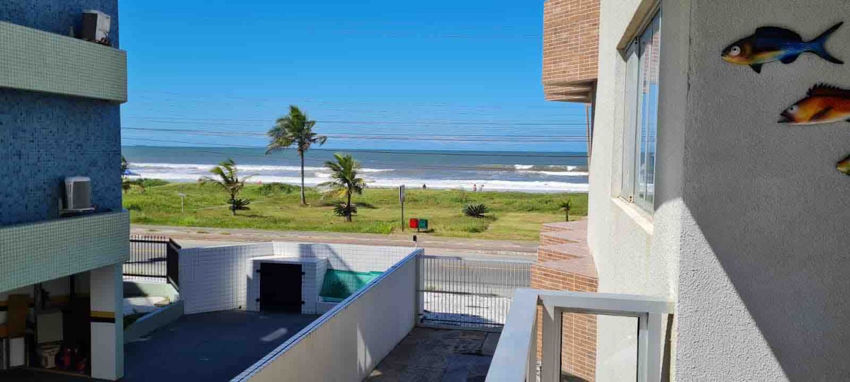 A seaside view is presented, showcasing the ocean waves gently lapping against the shore. Lush greenery and palm trees line the beach, with a clear sky above. The image captures a peaceful atmosphere, highlighting the proximity of the property to the waterside.
