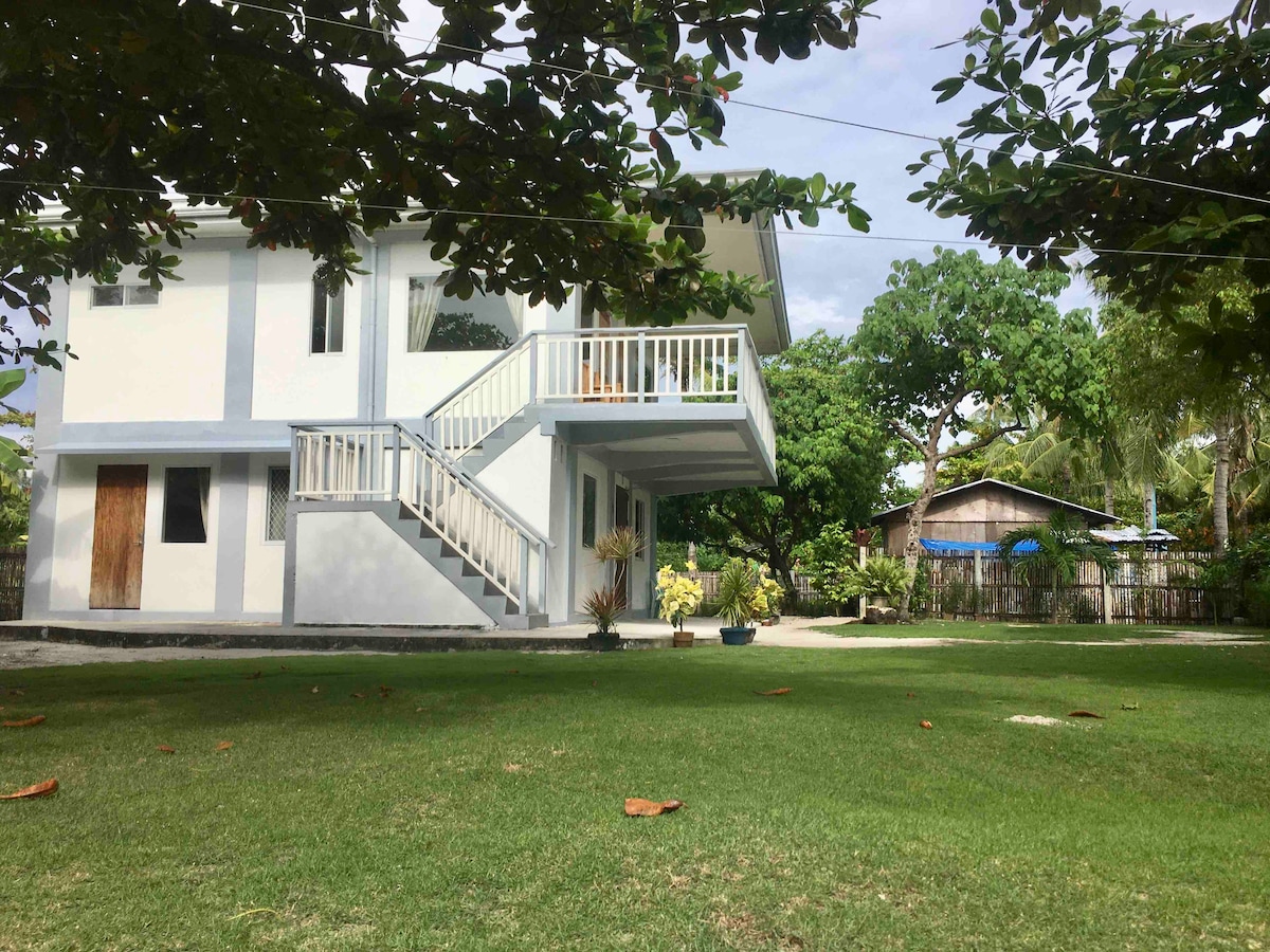 A two-story building is depicted, featuring a white exterior with blue accents. A staircase leads to a balcony that overlooks a spacious garden area. Surrounding greenery is abundant, creating a calm and inviting environment. Nearby, a local structure is visible through the trees.