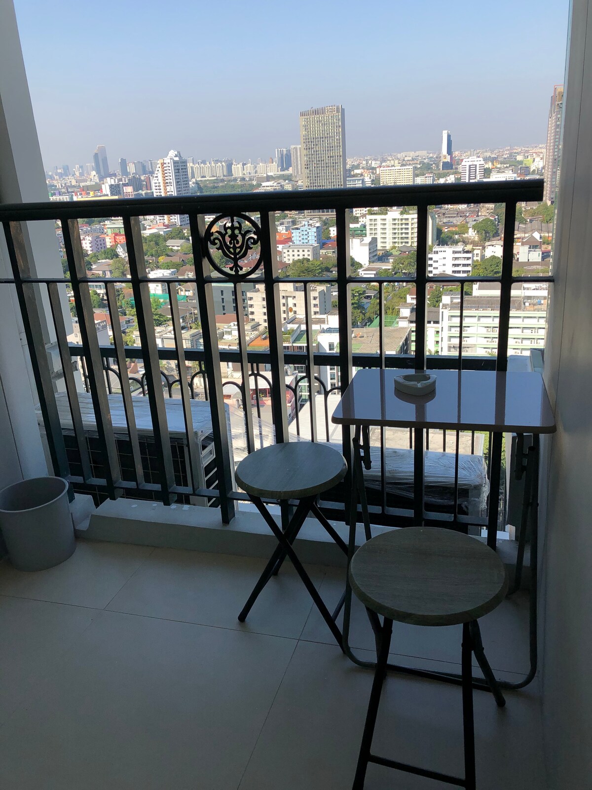 A balcony overlooks the Bangkok skyline, featuring two simple wooden stools and a small table. A metal railing encloses the space, while a waste bin is positioned nearby. The view captures a blend of city buildings and distant high-rises under a clear sky.