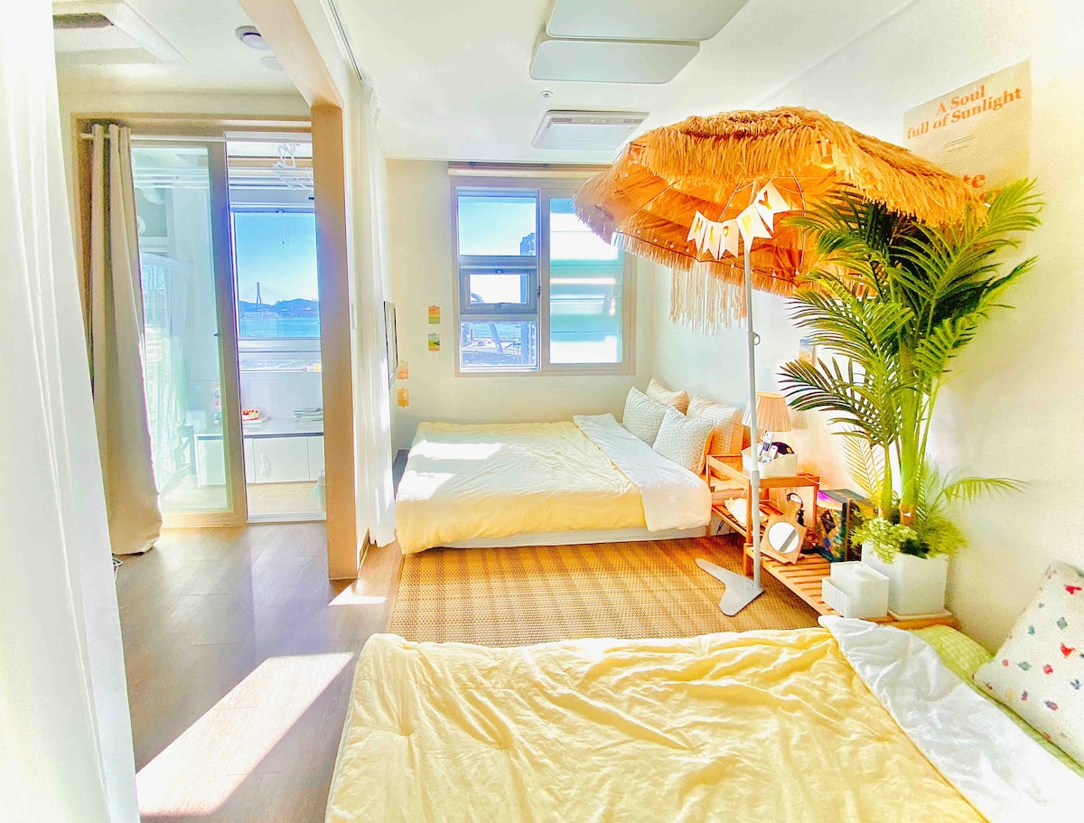 The room features two spacious beds adorned with soft bedding, complemented by a decorative straw umbrella and vibrant greenery. Natural light floods in through large windows, offering views of the ocean. A cozy mat lies on the floor, enhancing the inviting, open atmosphere.
