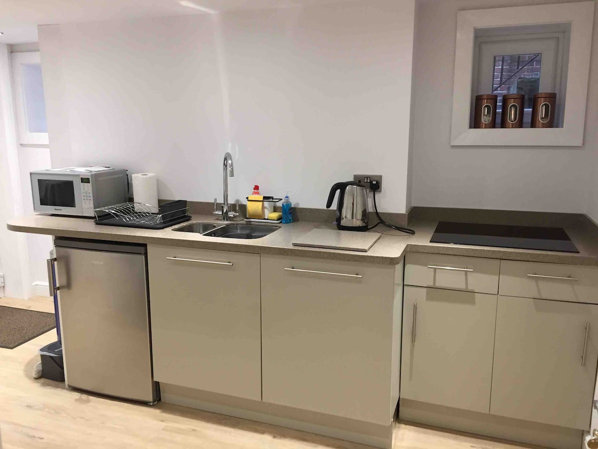 A modern kitchen area is equipped with essential appliances including a microwave, kettle, and refrigerator. A stainless steel sink and a stovetop are integrated into the sleek countertop, complemented by storage cabinets. Natural light is provided by a small window above the sink.