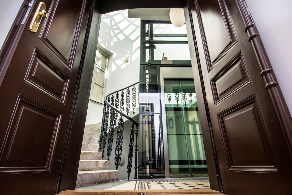 The entrance showcases a pair of grand wooden doors opening into a spacious lobby. A modern glass elevator is visible alongside an ornate staircase featuring decorative railings. Natural light filters through an overhead design, enhancing the contemporary and classic architecture.