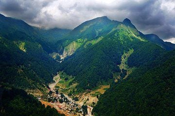 A panoramic view of rolling green mountains is presented, with deep valleys and scattered villages nestled among the trees. Thick clouds hover above, creating a dramatic contrast against the vibrant landscape, showcasing the natural beauty of the area.