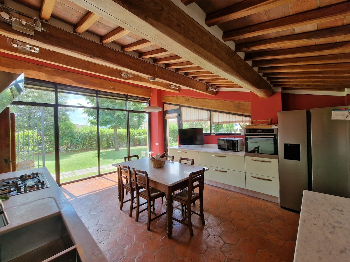A modern kitchen features a spacious dining table surrounded by wooden chairs. Large glass windows provide views of the garden, while an array of appliances are neatly arranged along the countertop. Exposed wooden beams add character to the ceiling.