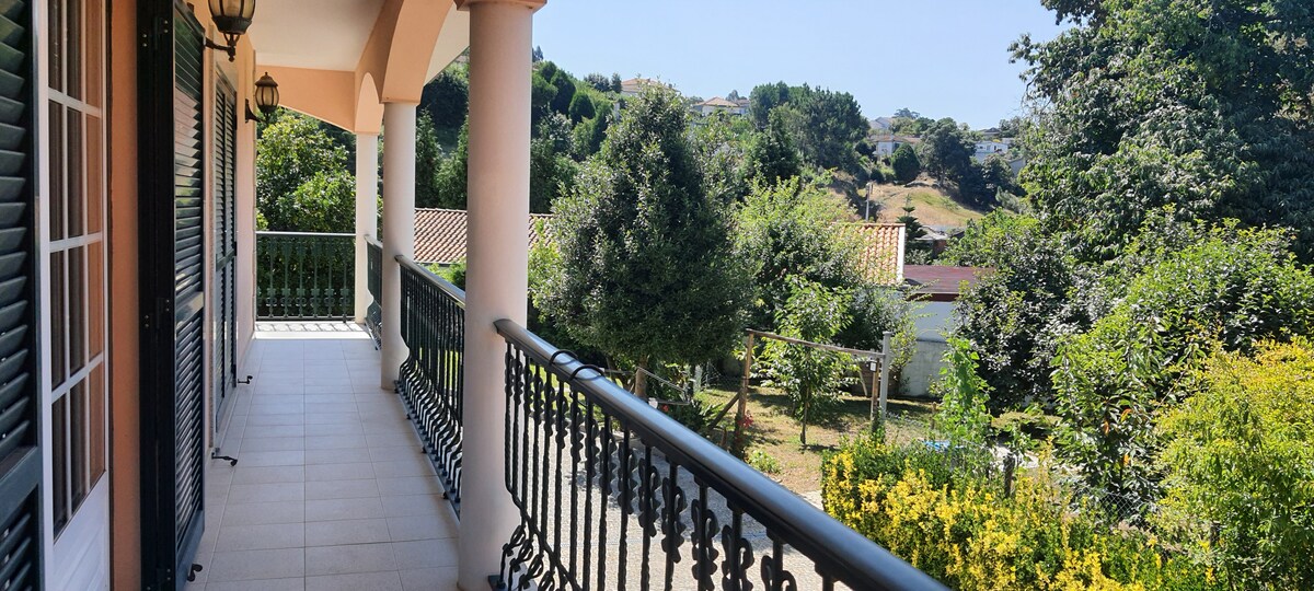 A spacious balcony features elegant railings, overlooking a lush garden with trees and flowering plants under clear skies. The surrounding hills are visible in the background, providing a serene natural setting.