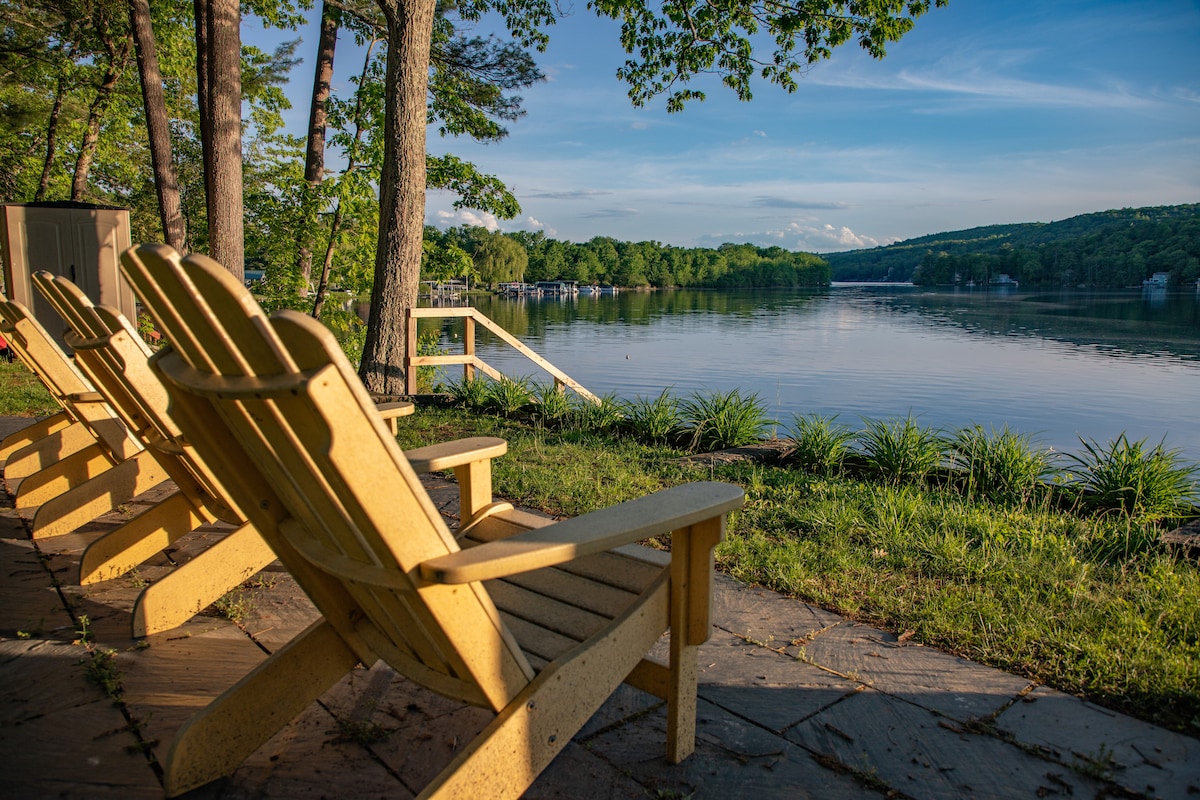 A row of Adirondack chairs is positioned on a stone patio, overlooking the calm waters of the lake. Lush greenery surrounds the area, and trees provide a natural border. The gentle slope leads to a wooden dock, enhancing the serene lakeside atmosphere.