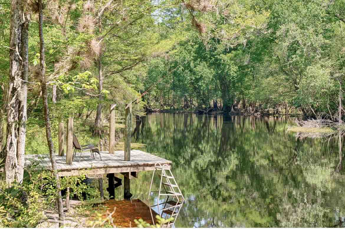 A serene dock extends over calm waters, surrounded by lush greenery and trees reflecting on the surface. A chair is positioned for relaxation, inviting moments of tranquility in nature.