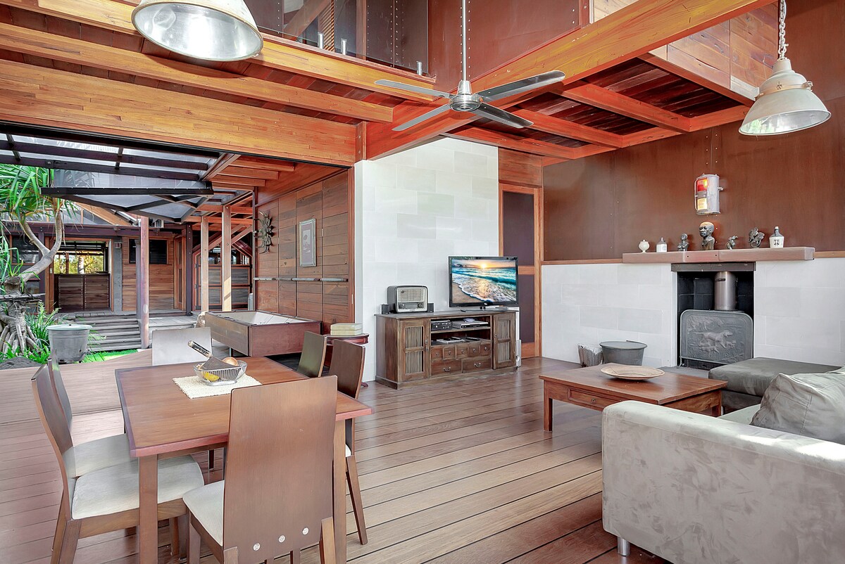 An open-concept living area showcases wooden floors and natural wood accents. A dining table is set for six, with a comfortable lounge area featuring a soft sofa. A flat-screen TV is mounted on a feature wall, while large sliding doors provide easy access to the outdoor courtyard.