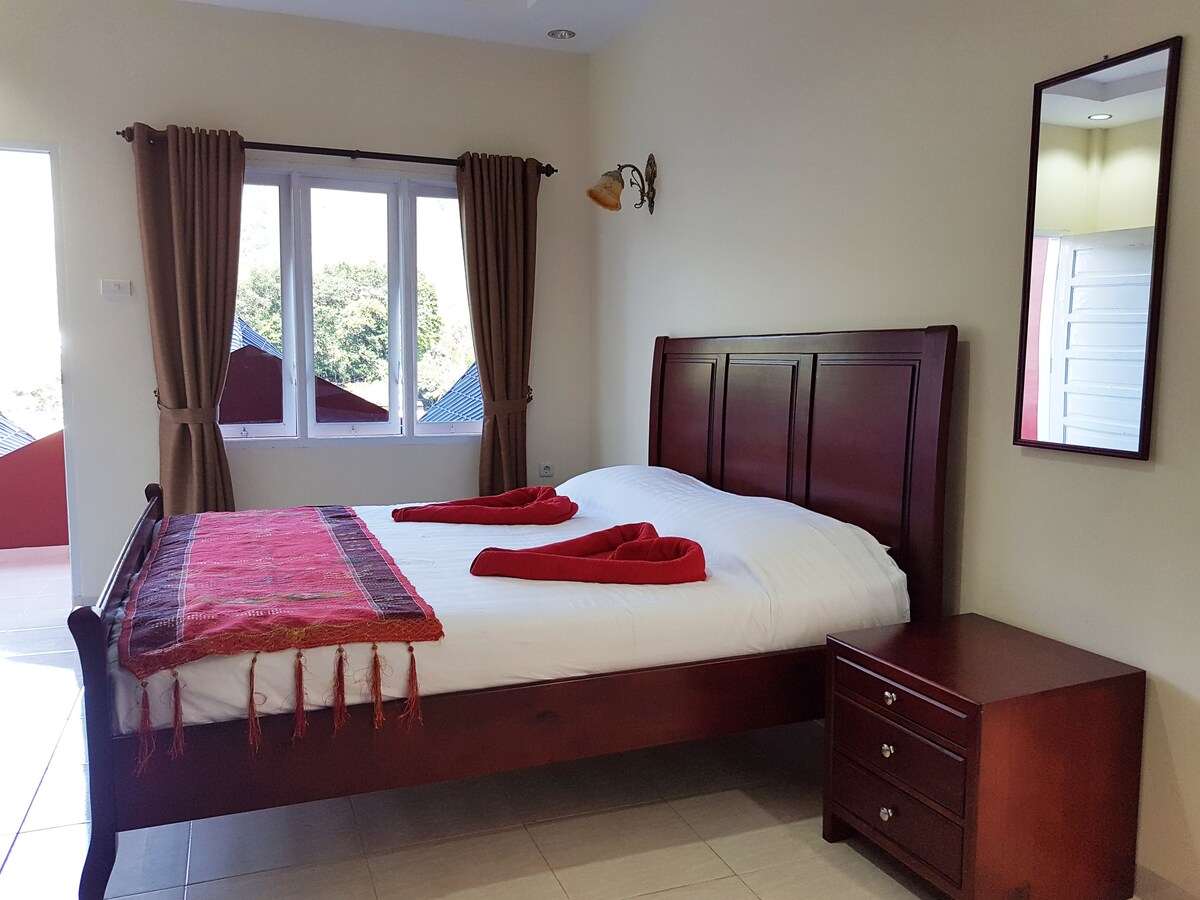 A cozy bedroom is highlighted by a dark wooden bed with crisp white linens and decorative red towels. A matching nightstand with drawers sits beside the bed. Natural light fills the room through multiple windows, complemented by soft curtain drapes.