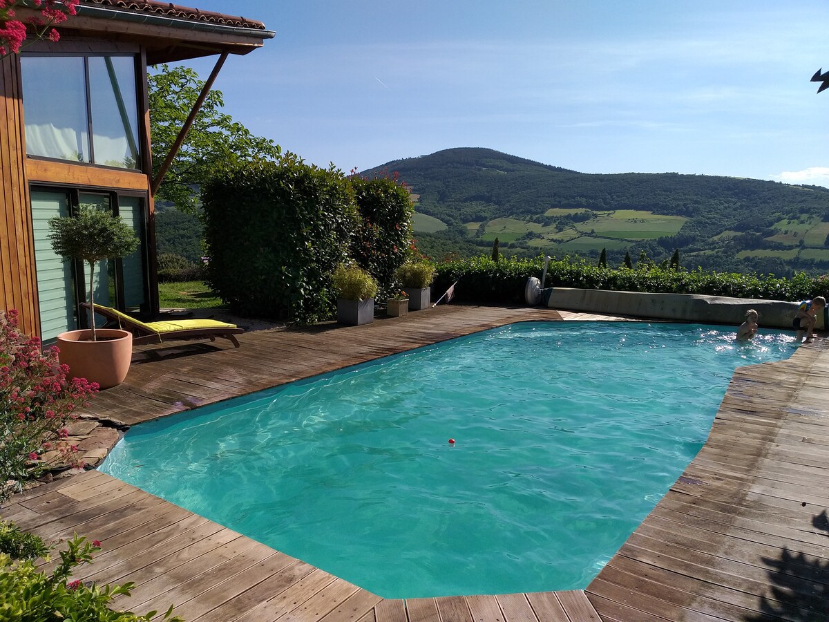 A refreshing pool is surrounded by wooden decking and landscaped greenery. Sun loungers are positioned nearby, while scenic hills rise in the background under a clear blue sky. Lush plant life adds a natural touch to the outdoor relaxation area.