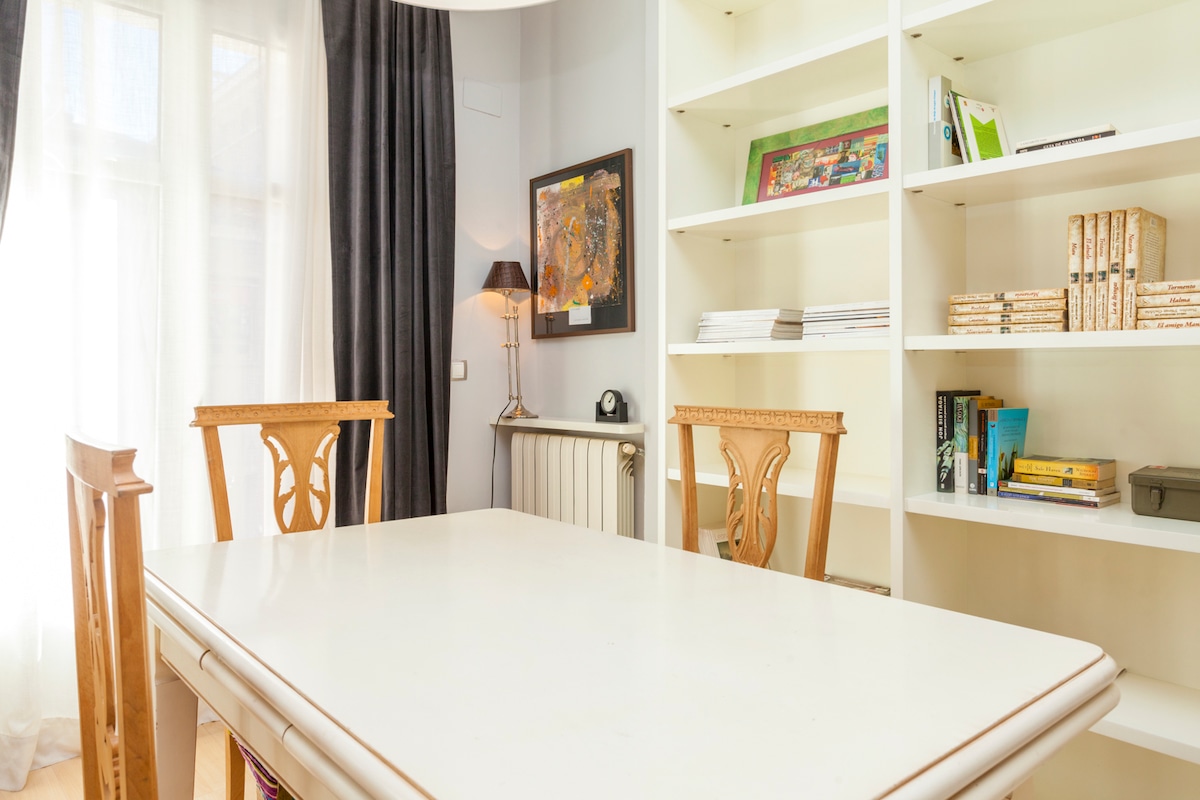 A bright dining area features a white table surrounded by four wooden chairs. A tall lamp stands beside the table, and a bookcase filled with various items is located against the wall. Natural light filters through sheer curtains, enhancing the room's inviting ambiance.