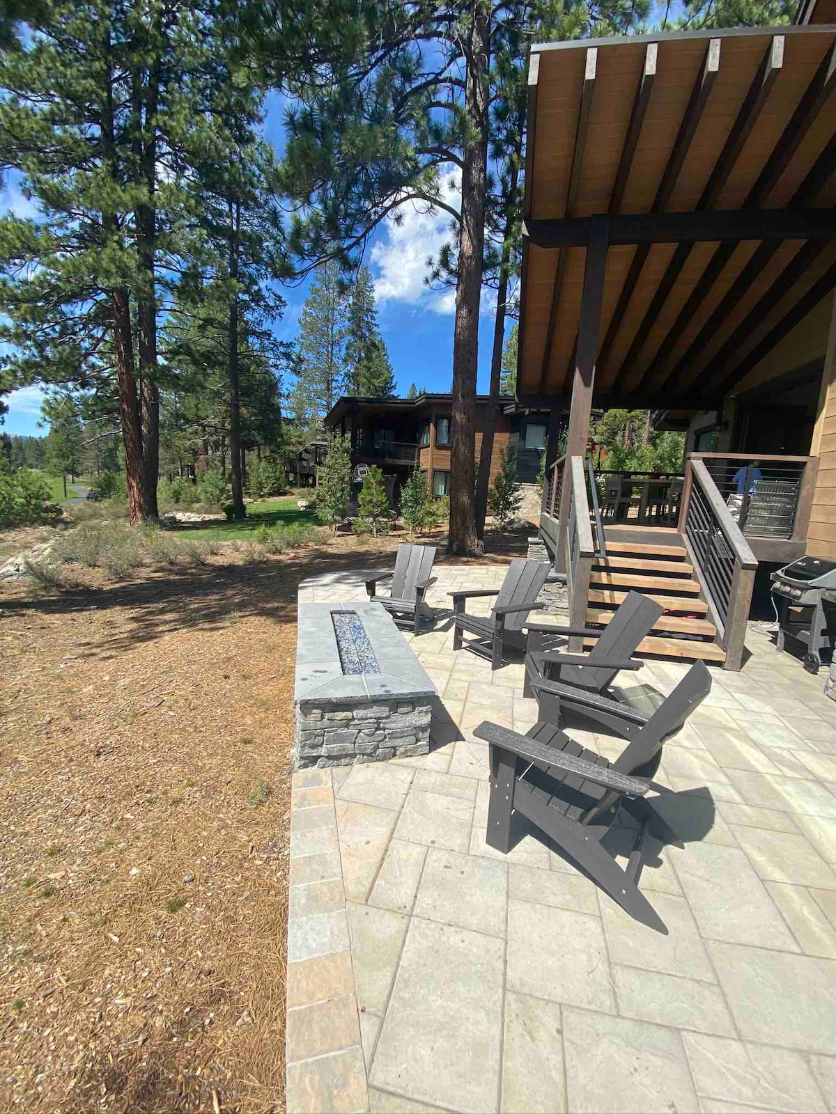 An outdoor patio features Adirondack chairs arranged around a stone fire pit, surrounded by natural landscaping. The backdrop includes tall pine trees and glimpses of neighboring homes. The space is designed for relaxation, inviting guests to enjoy the fresh air and tranquil surroundings.