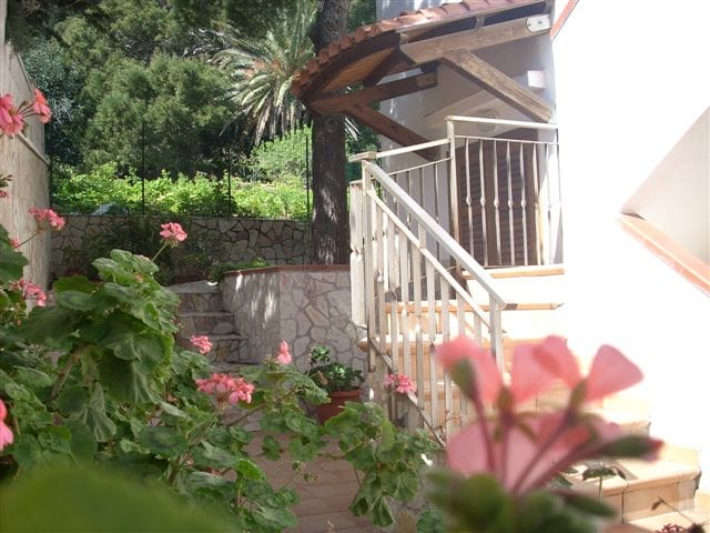 A staircase leads to a covered entrance, framed by decorative plants and a lush garden. Pink flowers are in the foreground, while tall trees and greenery provide a serene backdrop. Natural stone and wooden elements of the structure create a relaxed ambiance.