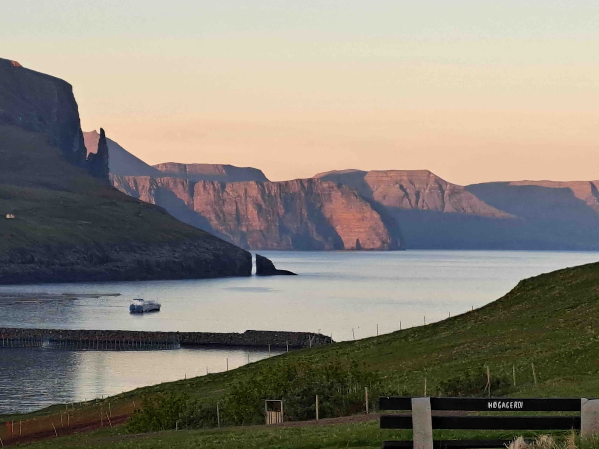 An expansive view of rugged cliffs rises above a calm sea, with a soft gradient of colors from the setting sun illuminating the landscape. In the foreground, green grass leads to a tranquil bay where a small boat is anchored near a rocky shoreline.