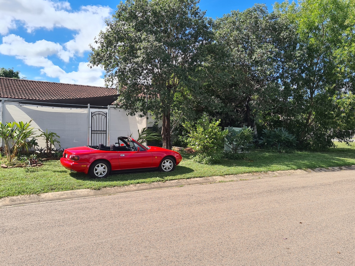 A vibrant red convertible is parked on the grass along a quiet street. Lush greenery is seen beside the vehicle, including trees and shrubs, while a white entry gate leads to the building in the background.