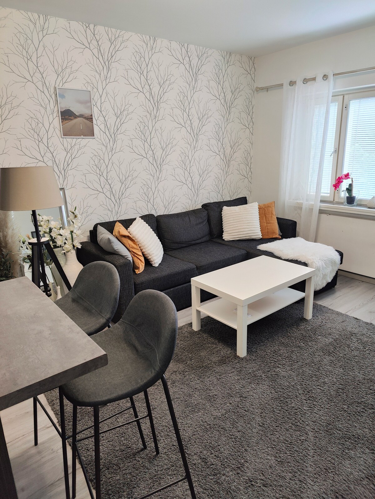 The living area features a dark gray sofa adorned with decorative pillows and a light throw. A small white coffee table rests on a textured area rug, with a bar table and two stools visible in the background. Natural light enters through the window, complemented by sheer curtains.