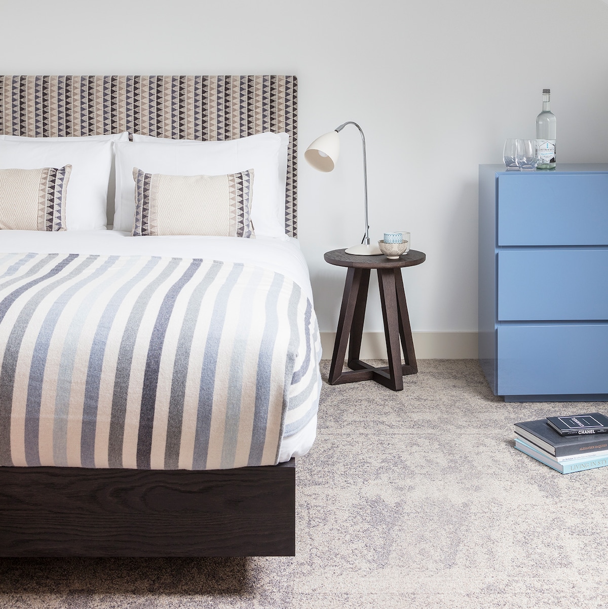 A comfortable sleeping area features a spacious bed adorned with a striped blanket. Stylish pillows complement the headboard, while a modern blue dresser offers additional storage. A small, dark wood side table holds a lamp, and the carpet adds a soft touch to the floor.