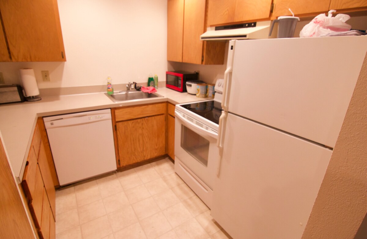 A compact kitchen features light wood cabinetry and a combination of white appliances, including a refrigerator, stove, and dishwasher. Counter space is available next to the sink, with a microwave and various kitchen items placed on the countertops. Neutral tiled flooring completes the space.