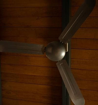 A ceiling fan with three metallic blades is mounted on a wooden ceiling. The design is sleek and modern, contributing to air circulation within the space. The warm wood tones create a cozy backdrop for the fan.