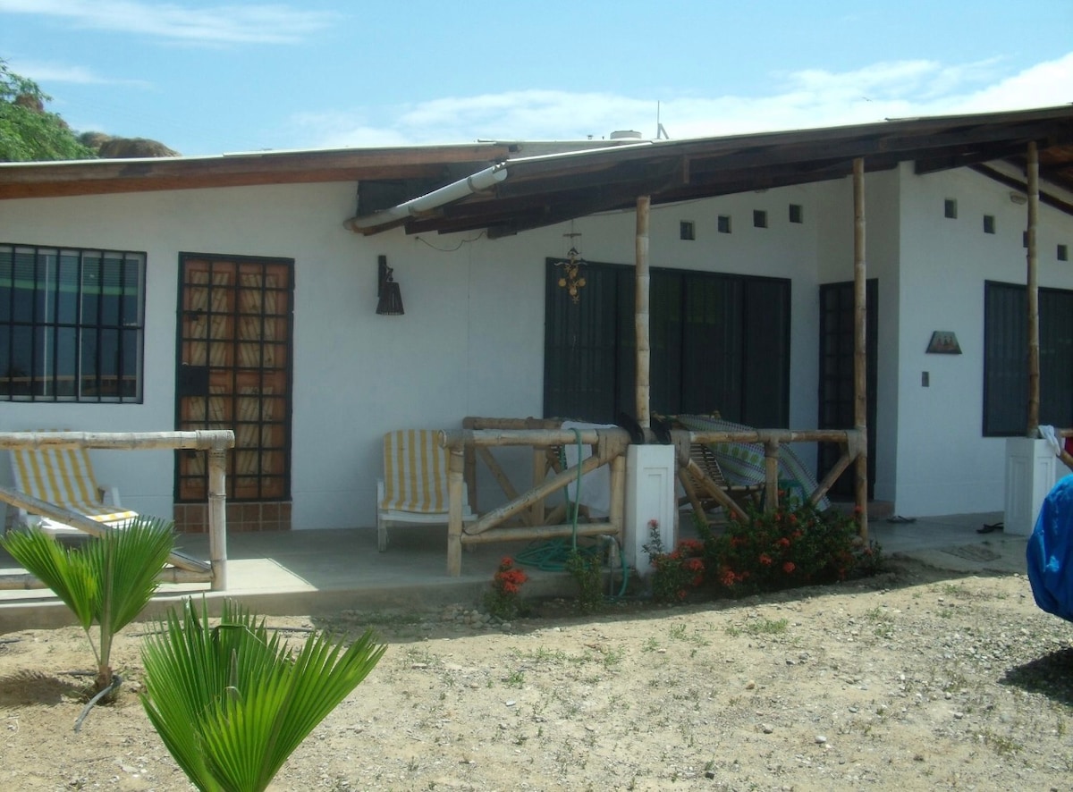 The exterior of the main house is depicted, featuring a covered porch with striped seating. Large windows provide natural light, and garden plants are visible in the foreground, enhancing the outdoor setting. The structure is surrounded by a gravel area under a clear sky.