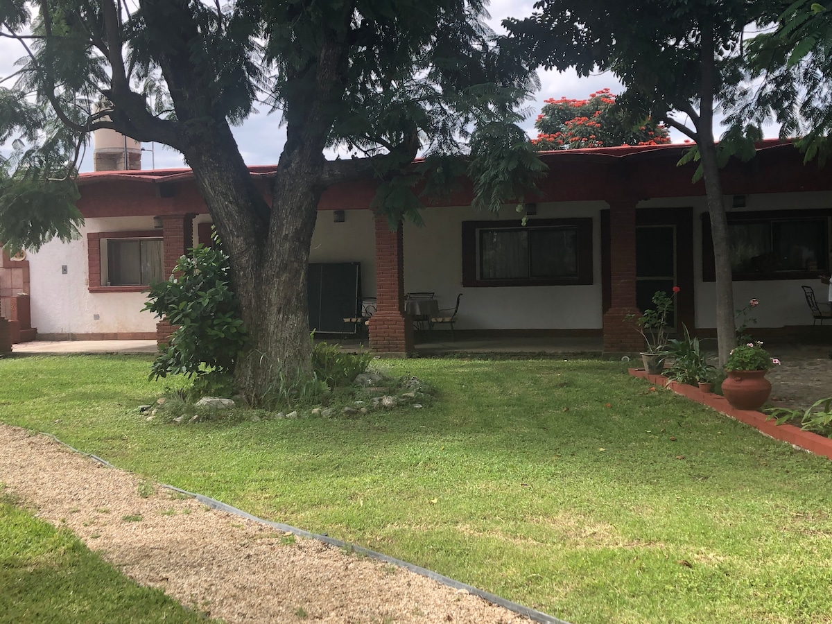 The exterior of the house is surrounded by a peaceful garden featuring green grass, a gravel path, and a large tree providing shade. The building has a rustic architectural style, with a red roof and several windows framed by dark green shutters.