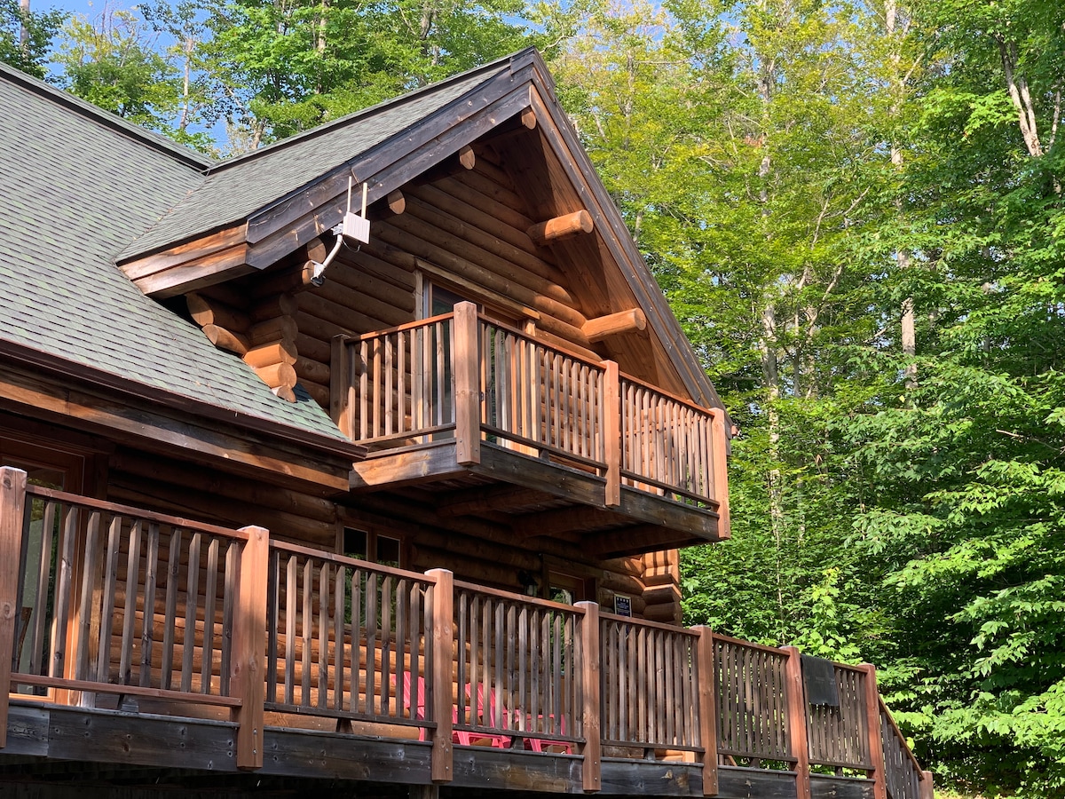The exterior of a log cabin is displayed, featuring a wraparound balcony with wooden railings. The natural wood construction is surrounded by lush green trees, indicating a forested setting. Sunlight casts a warm glow on the cabin's façade, highlighting its rustic charm.