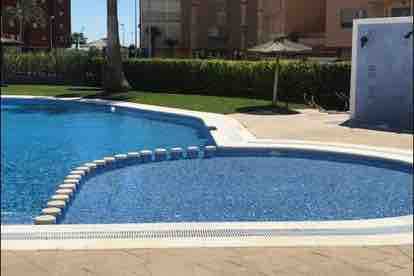 The image features a spacious swimming pool with vibrant blue tiles, surrounded by landscaped greenery. A shallow area is visible, designed for relaxation, and a sunshade is positioned nearby, enhancing the poolside setting. The edge of the pool is lined with white tiles.