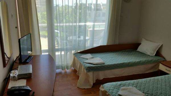 A room features two single beds dressed in light blue covers, positioned beside a wooden desk and a television. Sheer curtains frame a large window, allowing natural light to fill the space. A balcony is visible outside, suggesting potential outdoor seating.