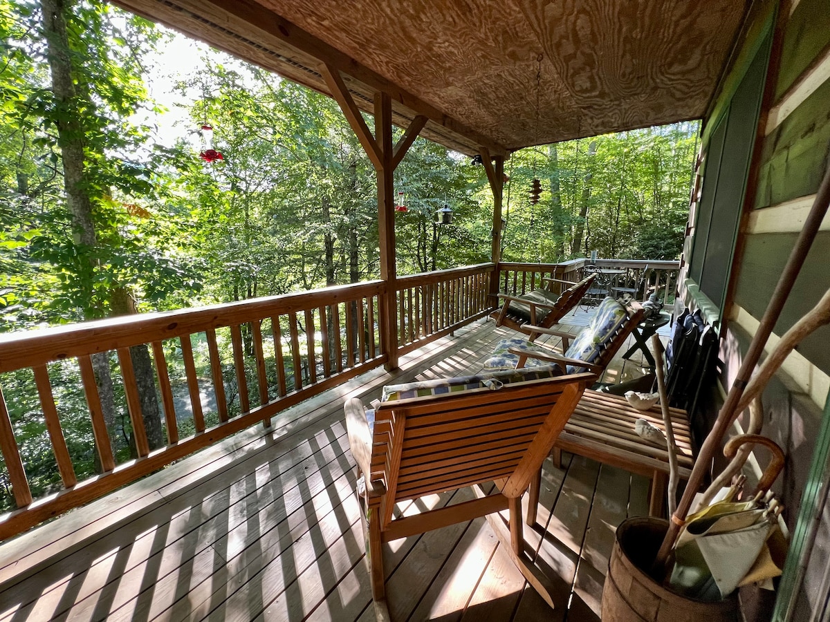 The wraparound porch features wooden seating with a rustic design, surrounded by lush greenery. Sunlight filters through the trees, casting gentle shadows on the wooden deck. Bird feeders and nature-inspired decor add to the serene atmosphere of this tranquil outdoor space.