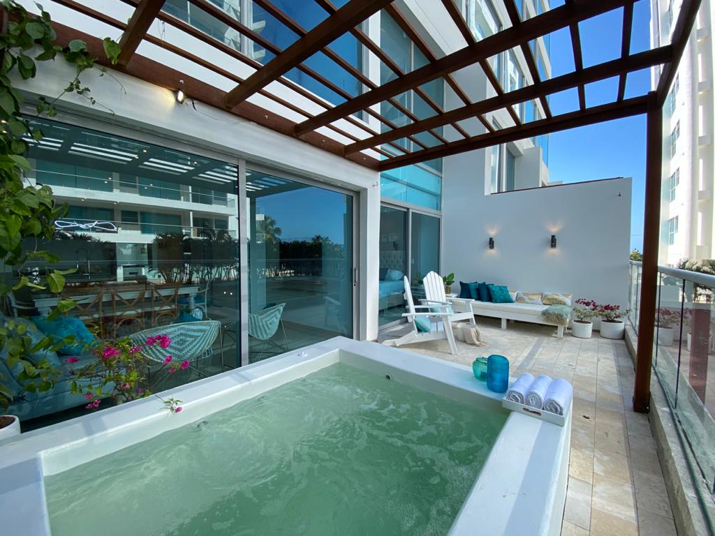 A private jacuzzi is situated on a covered terrace, surrounded by greenery. Large glass doors provide a view of the outdoor space, which is furnished with a comfortable seating area. Natural light fills the area, contributing to a relaxing ambiance.