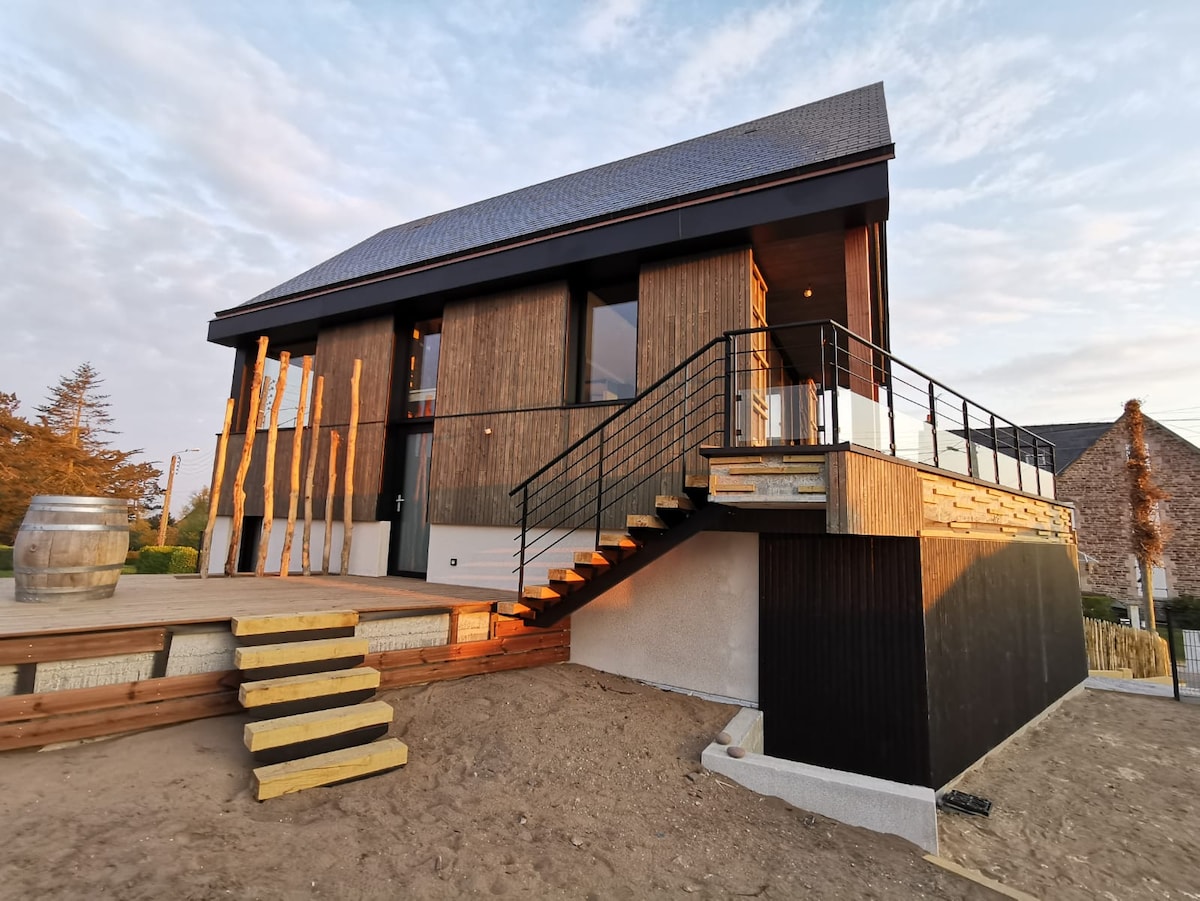 A modern building features a combination of wooden and sleek materials, with large windows allowing natural light. Steps lead up to a spacious terrace, complemented by railings. The surrounding area is characterized by a sandy surface, indicating an outdoor setting.