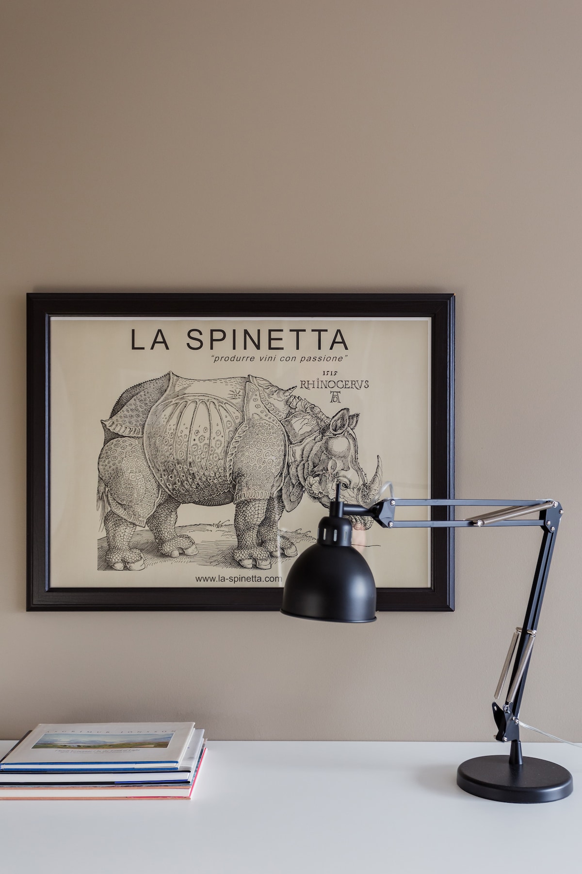 A framed vintage poster of a rhinoceros adorns the wall, accompanied by a modern black desk lamp. A few stacked books sit on the white tabletop, creating an organized and calming workspace.