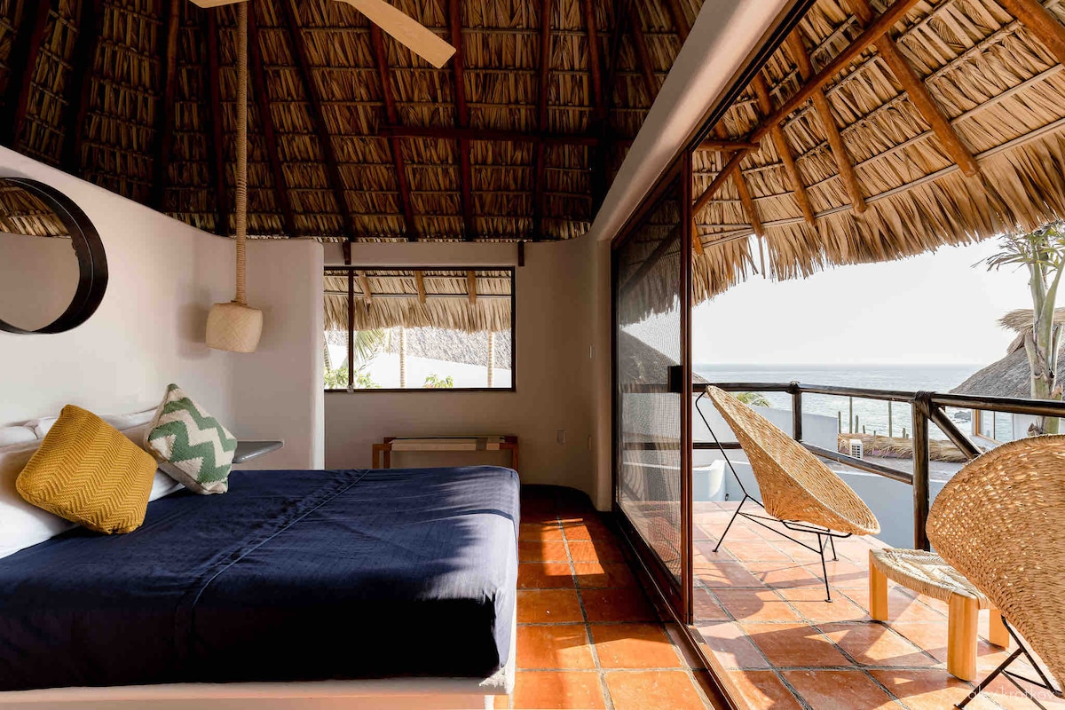 A master suite features a spacious bed with a dark blue coverlet and decorative pillow. Large glass doors open up to a private terrace, allowing natural light to fill the room. A straw palapa roof creates a relaxed atmosphere, while inviting views of the surrounding area are visible.